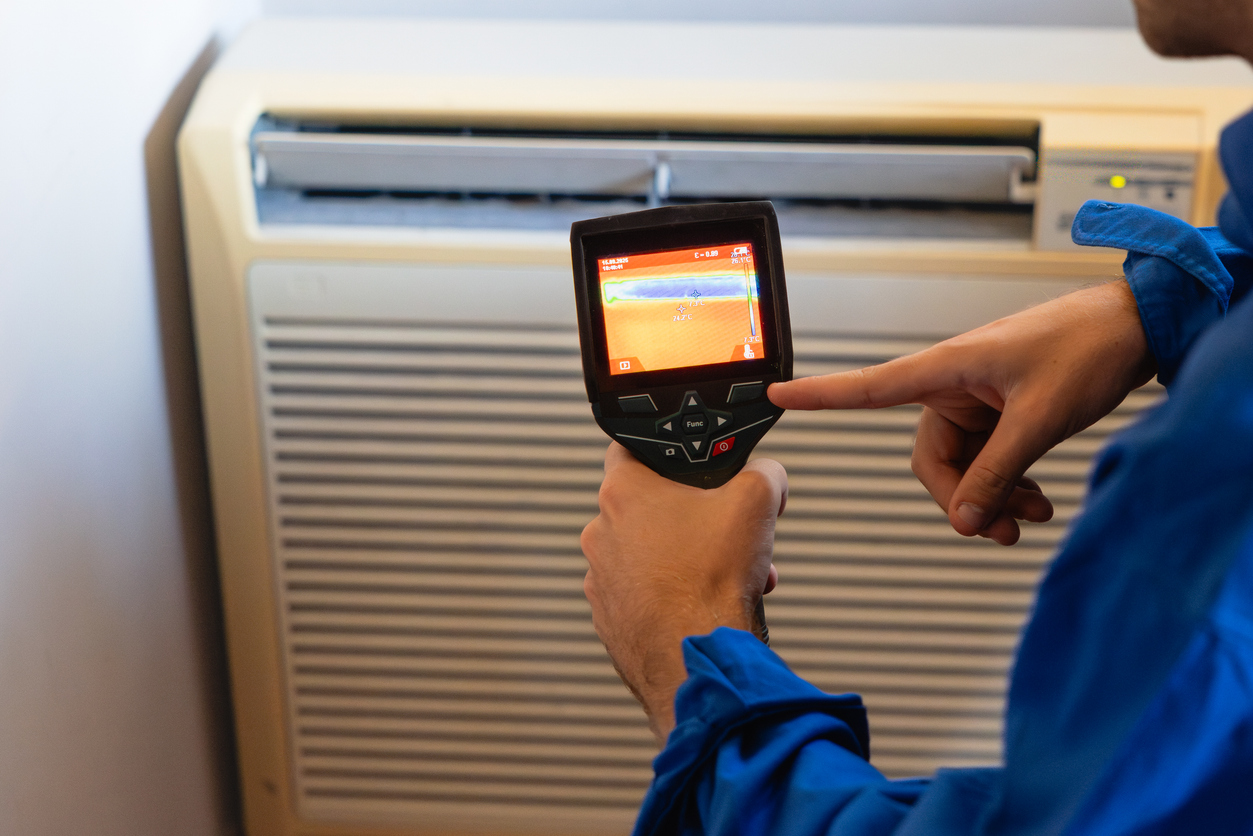 A technician using a thermal imaging camera to detect air leaks near an air conditioning unit.