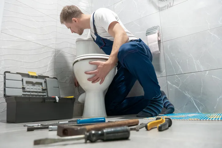 A plumber is kneeling beside a toilet in a bathroom, performing repairs.