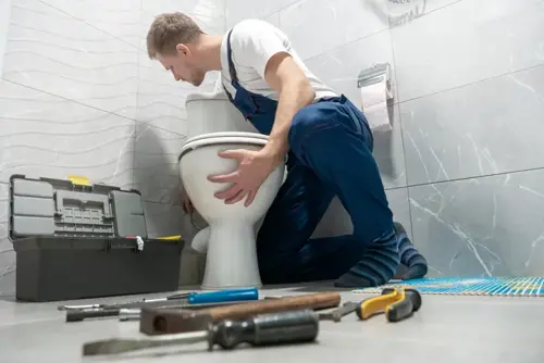 A plumber is kneeling beside a toilet in a bathroom, performing repairs.
